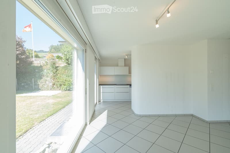 Bright, unfurnished room with white tiled flooring, white walls, and large windows offering a view of a green outdoor area. The room features modern lighting fixtures and a small, white kitchen area with cabinets and a countertop.