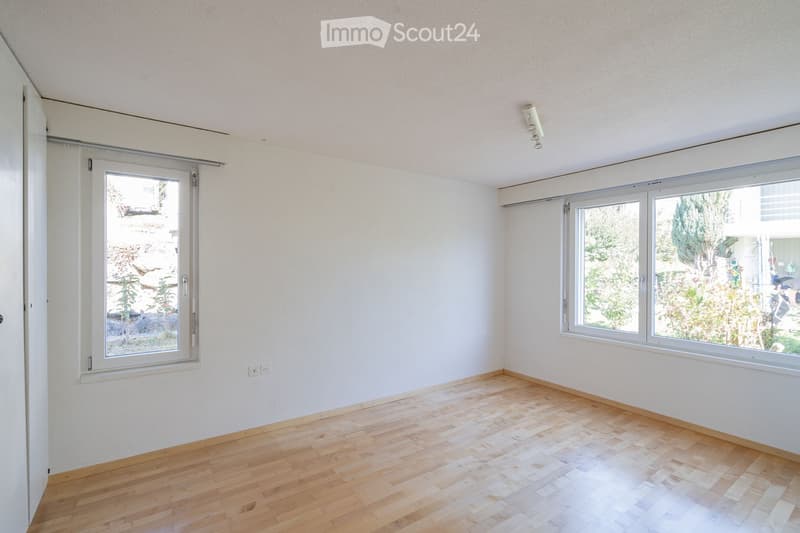 Empty room with wooden flooring, white walls, and large windows offering views of greenery and a street.