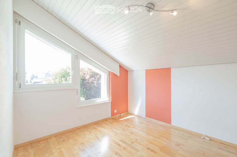 Empty room with wooden flooring, white walls, and large windows. The ceiling has a modern design with built-in lighting. The walls feature a two-tone color scheme with white and orange sections.