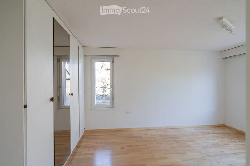 Empty room with light wooden flooring, white walls, and large windows allowing natural light. The room features a sliding door leading to another space and a visible air conditioning unit on the wall.