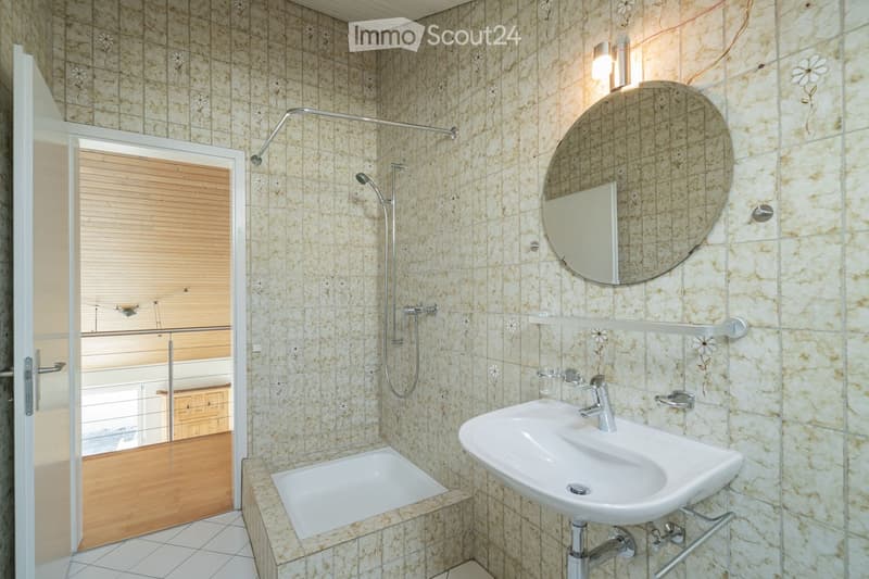 Bathroom with beige tiled walls, a white sink, a bathtub, a large round mirror, and a shower with a handheld sprayer. The room features a wooden door with blinds and a small shelf above the sink.