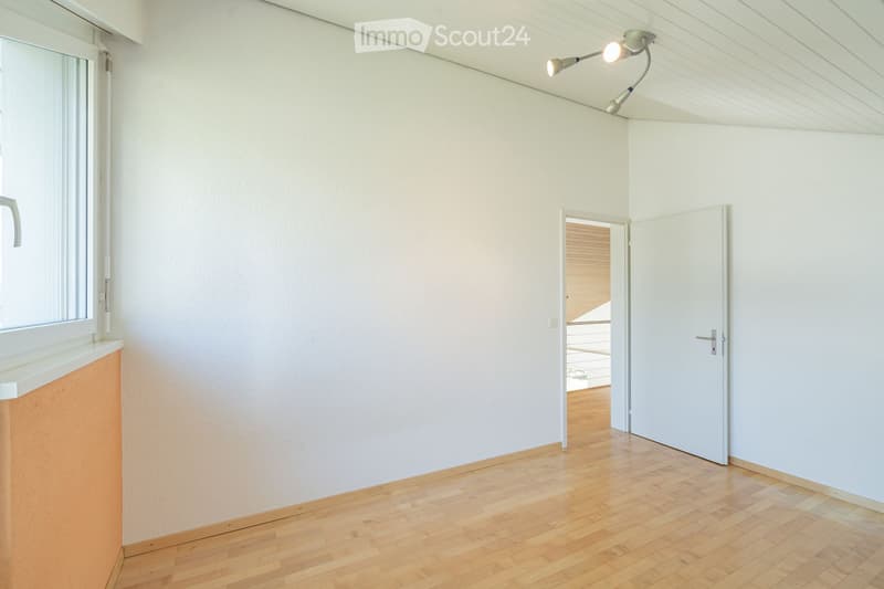 Empty room with light-colored walls, wooden flooring, and a large window with blinds. A door leads to another room with wooden flooring and a visible balcony area.