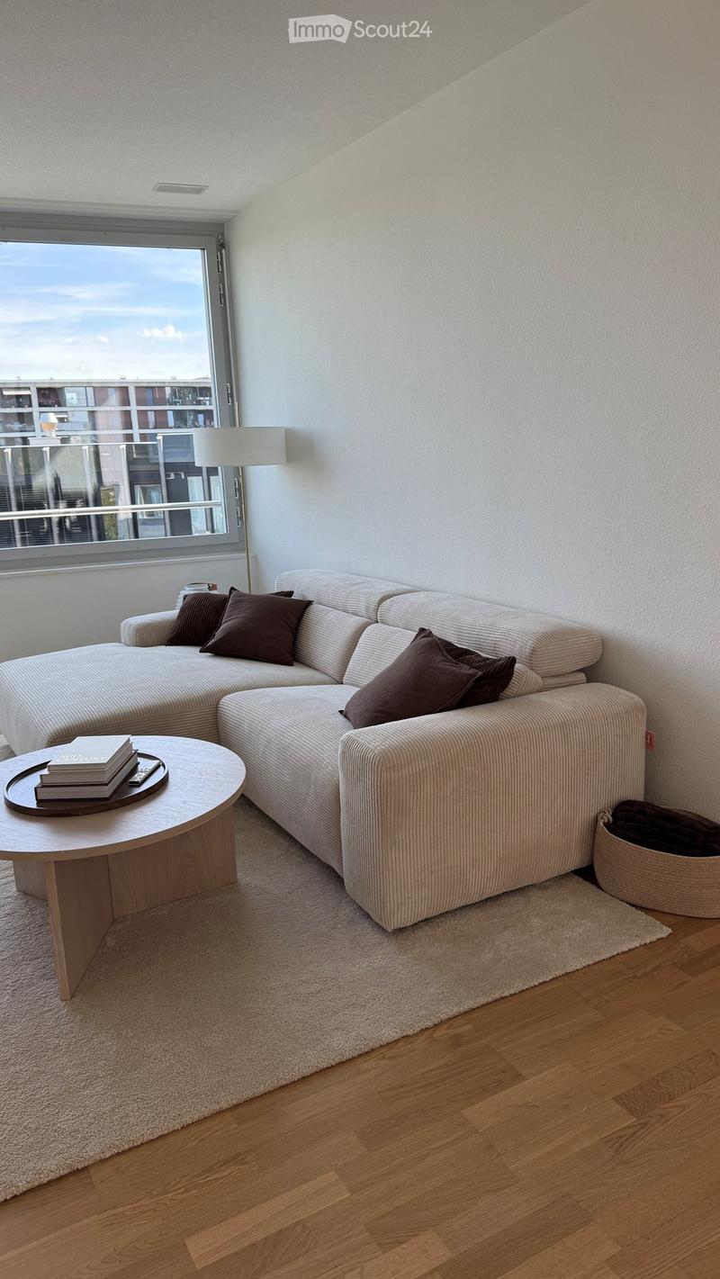 Modern living room with a white sectional sofa, wooden coffee table, books, and a floor lamp near a large window offering a view.