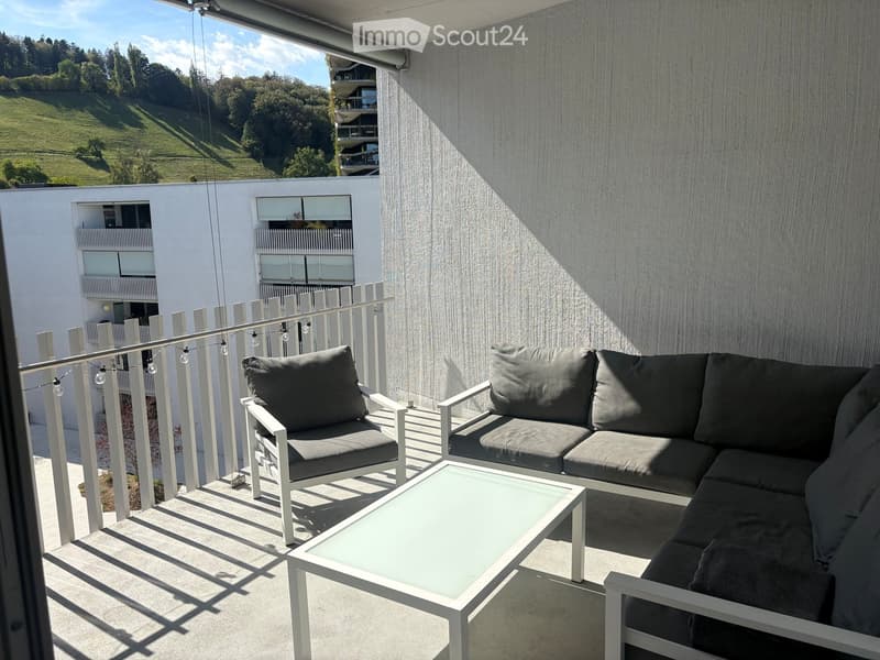 Balcony with modern white furniture, including a sofa and a glass coffee table, overlooking a green hillside with trees and modern buildings.