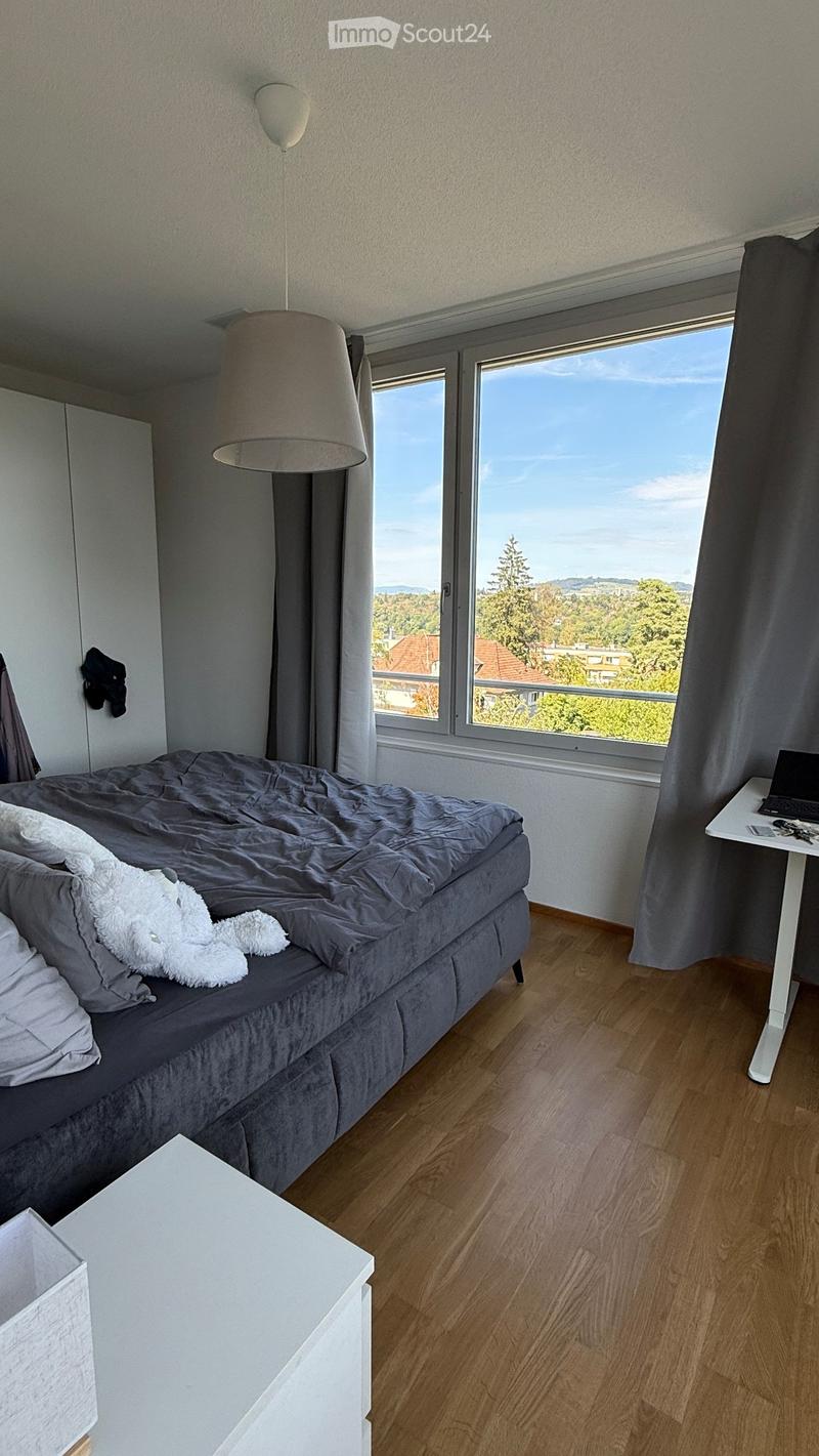 Modern bedroom with a grey duvet, white pillows, a white fluffy throw, a white side table, a laptop on a white desk, and large windows offering a scenic view.