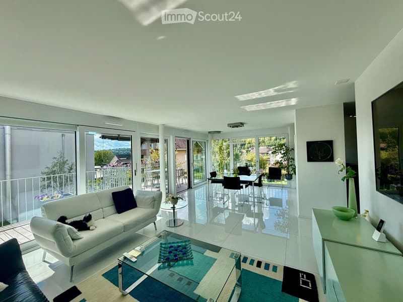 Modern living room with white walls, large windows, and a balcony. Features a white sofa, coffee table, dining area with chairs and table, and a TV screen.