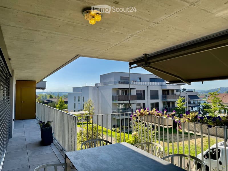 Balcony with a table and chairs, overlooking a modern residential complex with greenery and a clear blue sky.