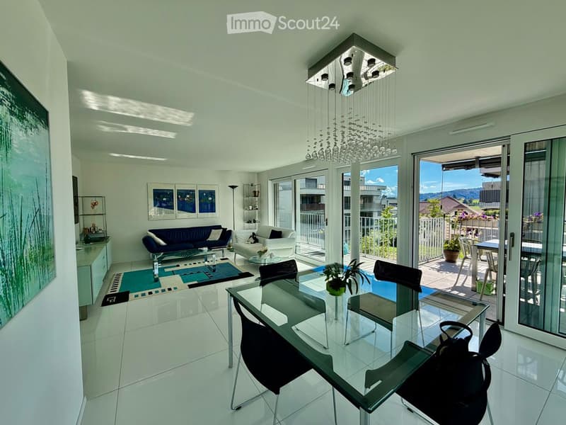 Modern living and dining area with white walls, large windows, and a balcony. Features a blue sofa, white armchair, glass coffee table, glass dining table with black chairs, and a chandelier.