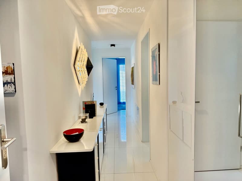 Modern hallway with white walls, tiled floor, and recessed lighting. Features a small console table with decorative items and framed artwork on the walls.