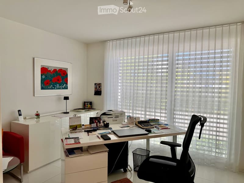 Modern office space with a large desk, ergonomic chair, laptop, printer, calendar, picture frame, and a small lamp. Large windows with sheer curtains provide natural light.