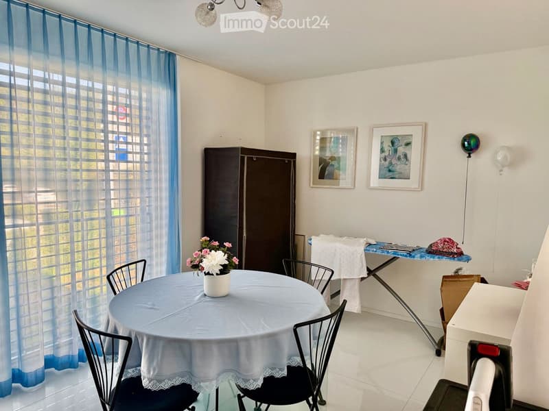 A furnished room with a round table, chairs, a flower vase, a large window with blue curtains, a picture frame, a cabinet, and an ironing board.