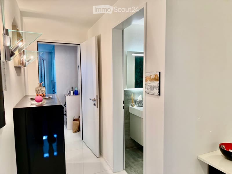 Modern hallway with white walls, tiled floor, and glass wall-mounted shelves. An open door leads to a bathroom with a sink, mirror, and decorative items.