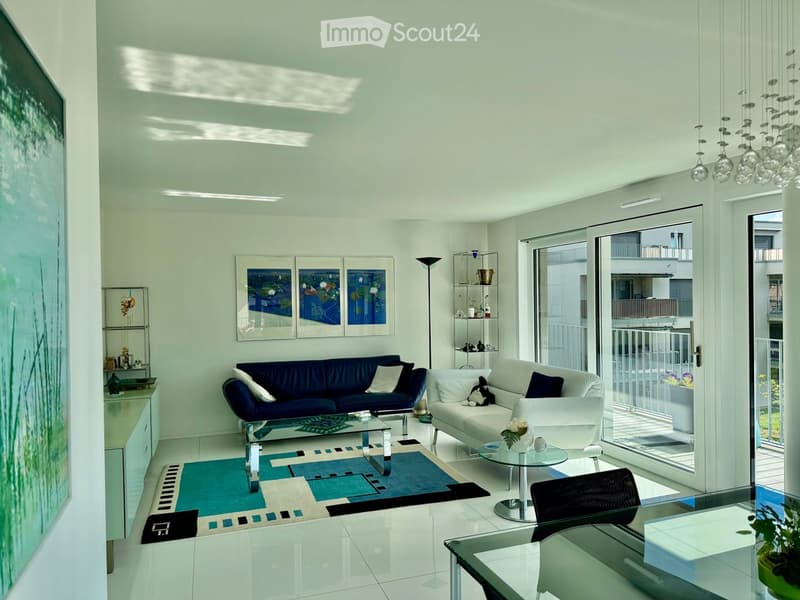Modern living room with white and blue color scheme, large windows, balcony access, sofa, coffee table, chairs, shelves, and artwork.