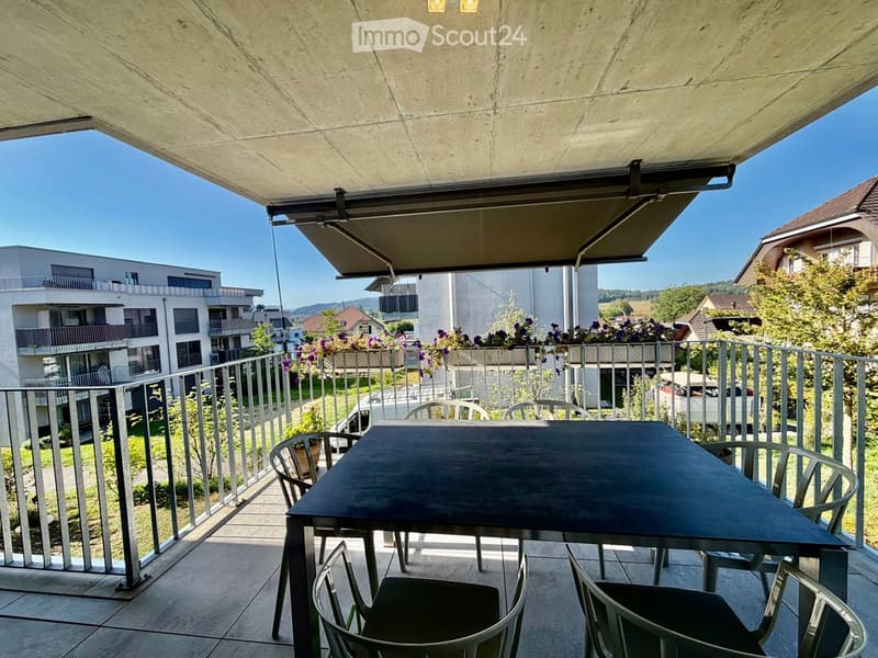Balcony with a table and chairs, metal railing, and a retractable awning, overlooking a residential area with modern buildings and greenery.