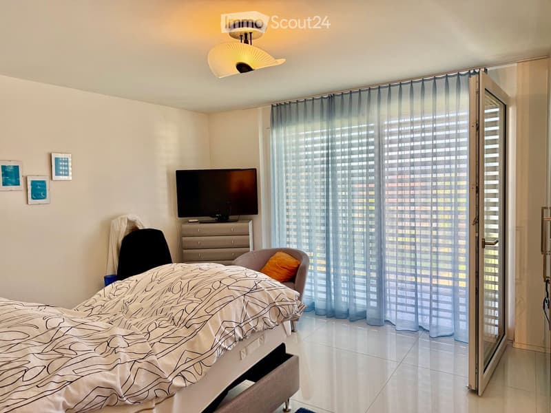 Modern bedroom with a large bed, a television on a dresser, a chair with an orange pillow, and a large window with sheer curtains. The room features a sleek, contemporary design with light-colored walls and a tiled floor.