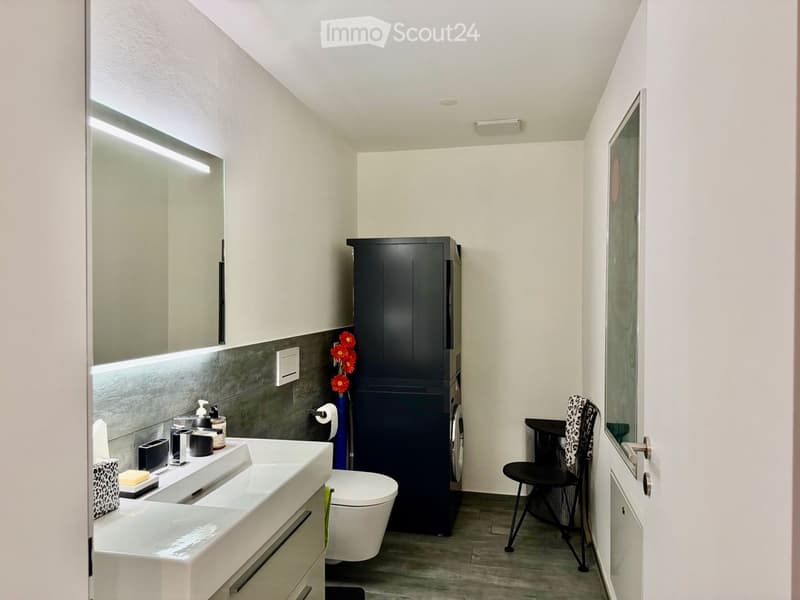 Modern bathroom with a white sink, toilet, large mirror, dark cabinet, and a small chair near the door.