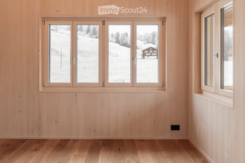 Empty room with light wooden walls and floor, large windows showing a snowy outdoor view.