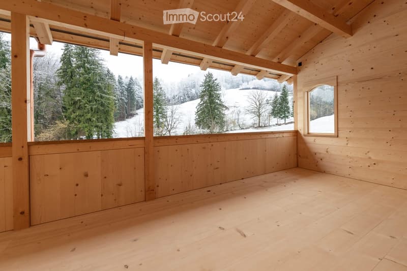 Empty wooden room with exposed beams, large windows overlooking a snowy landscape, and a small arched window on the right side.