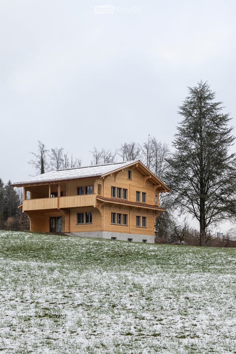 A two-story wooden house with a sloped roof and large windows, situated on a grassy, snow-covered hill.
