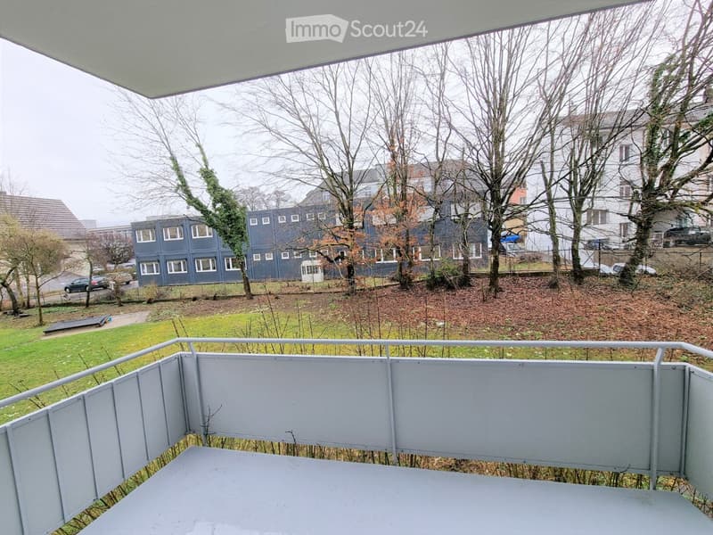 Balcony with grey railing and table, overlooking a grassy area with trees and a multi-story building in the background.