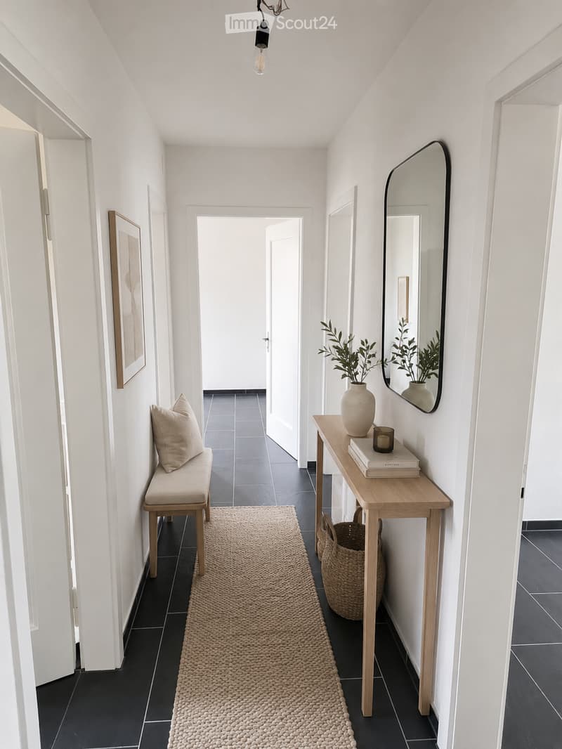 A modern hallway with white walls, dark tiled flooring, and a beige runner rug. Features include a wooden bench with a cushion, a small wooden table with a vase, books, and a candle, a large mirror, and a woven basket on the floor.