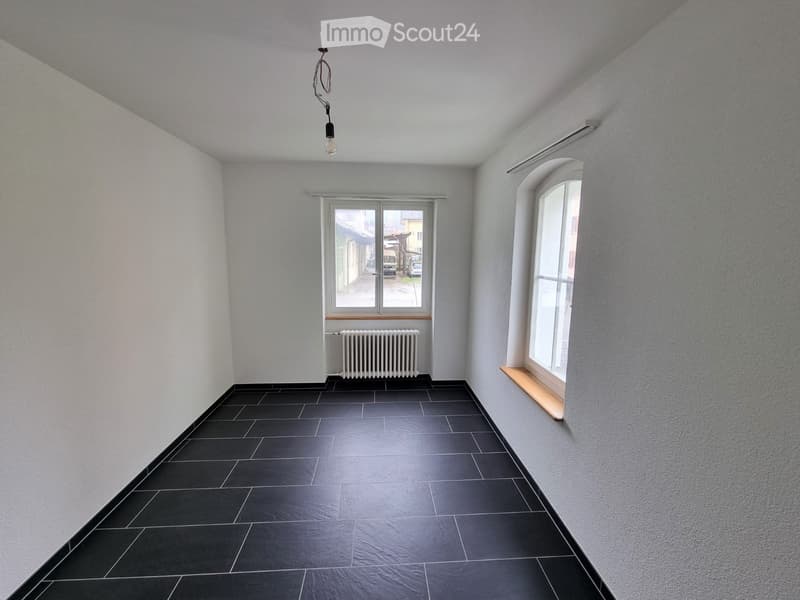 Empty room with light grey walls, black tiled floor, and a single window with a radiator below it. The room has a simple, modern design with a pendant light fixture hanging from the ceiling.