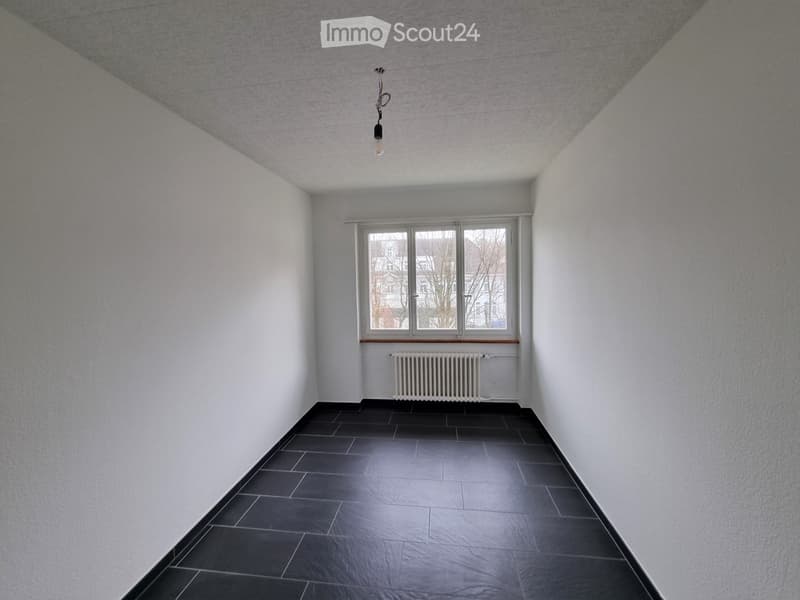 Empty room with white walls, dark tiled floor, single window with radiator below, and a single light fixture on the ceiling.