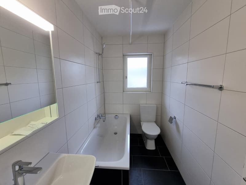 Modern bathroom with white tiles, bathtub, toilet, sink, mirror, and showerhead. Features a window and built-in towel rack.