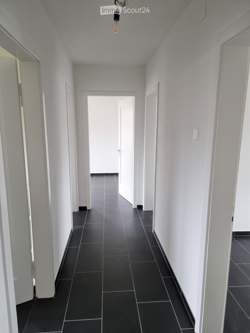 A long, narrow hallway with white walls and black tiled flooring. The hallway features several doors on both sides and a single light fixture hanging from the ceiling.