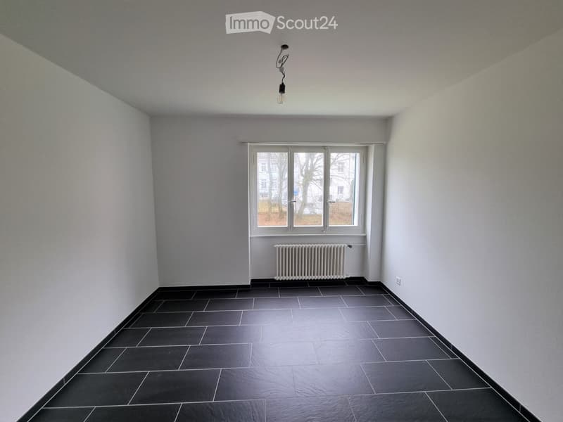 Empty room with grey walls, black tiled floor, a window with white frame, radiator, and a single light fixture on the ceiling.