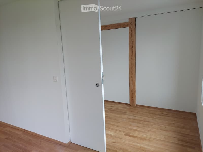 Empty room with light wooden flooring and white walls, featuring a closed white door and a visible electrical outlet.