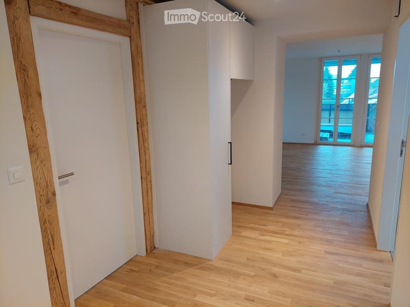 Empty hallway with wooden flooring, white walls, and a door leading to a room with large windows and a balcony view.