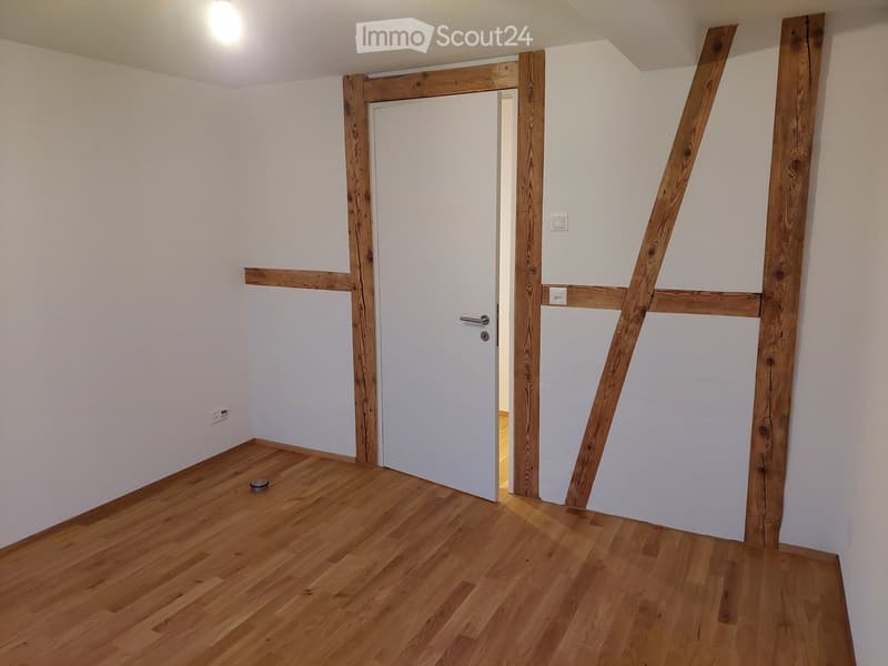 Empty room with wooden flooring, white walls, and a white door leading to another space. Exposed wooden beams frame the doorway.