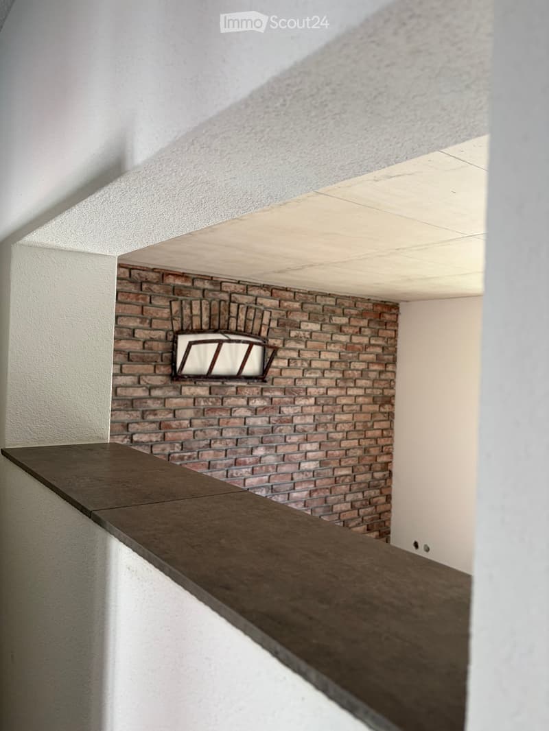 A narrow hallway with a brick wall, a small window, and a wooden railing leading to an open space.
