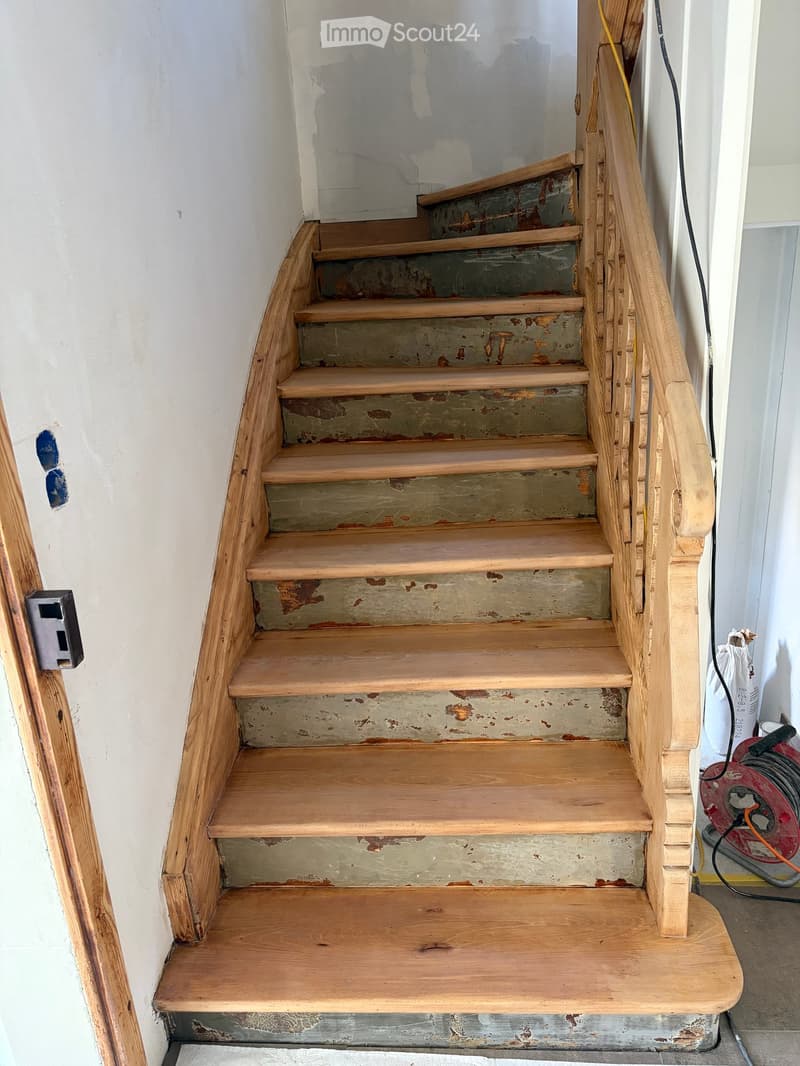 Wooden staircase with a handrail, showing a mix of polished and unpainted steps, and a visible electrical outlet on the wall.