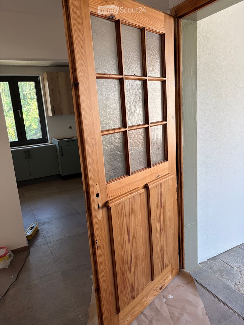 Empty hallway with a wooden door featuring frosted glass panels, tiled floor, and visible kitchen area with cabinets and a window in the background.
