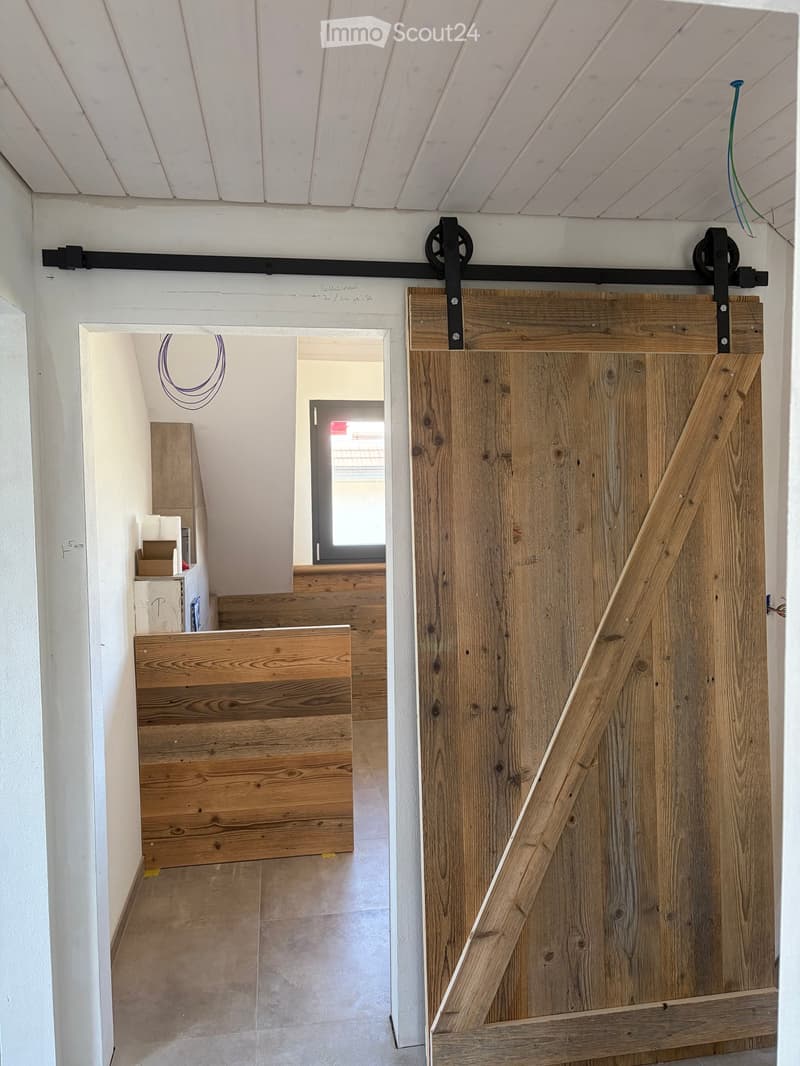A hallway with a large wooden sliding door on the right and a partially open doorway leading to another room on the left. The walls are white, and the floor is tiled.