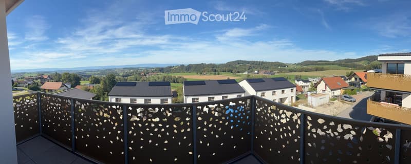 Balcony with decorative metal railing, overlooking a scenic countryside with houses, fields, and distant hills under a clear blue sky.
