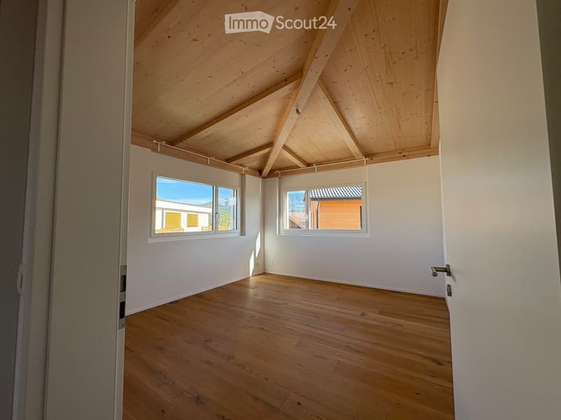Empty room with wooden floor, white walls, and exposed wooden ceiling beams. Two windows offer natural light and views of the outside.