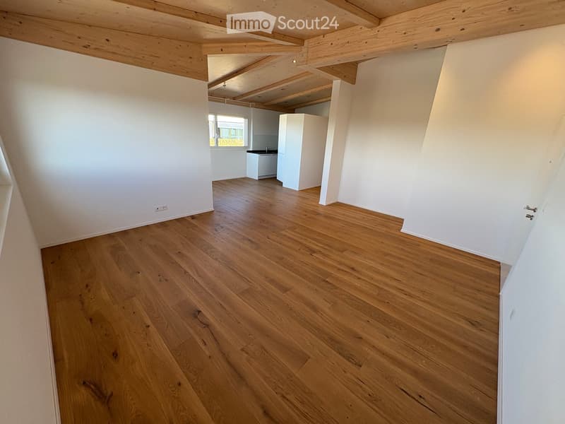 Empty room with wooden flooring, white walls, and exposed wooden beams on the ceiling. A small kitchen area is visible in the background with a sink and cabinets.