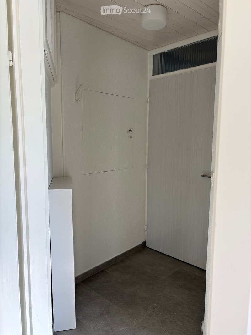 Empty hallway with white walls, tiled floor, and a closed door at the end. A small white shelf is visible on the left side.