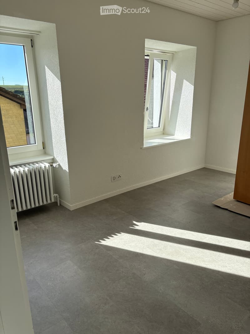 Empty room with tiled floor, white walls, and two windows allowing natural light. A radiator is visible on the left side.