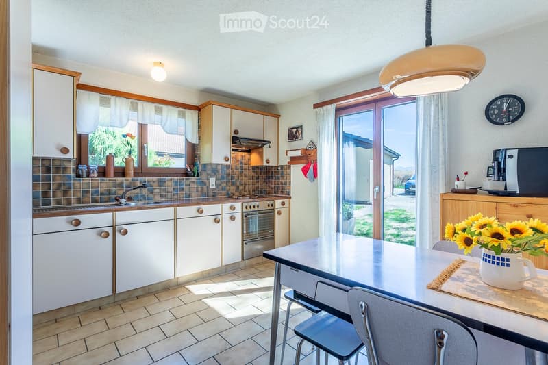 Bright kitchen with white cabinets, a central island table, and a dining area. Features include a sink, oven, and a large window with a view of the outdoors.