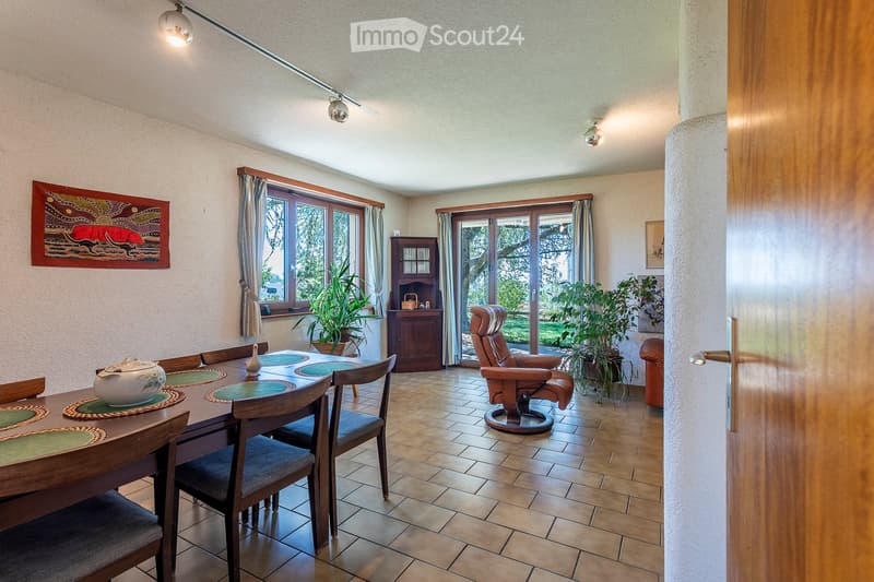 Dining room with a wooden dining table, chairs, a teapot, a recliner, and plants near large windows and a sliding glass door.