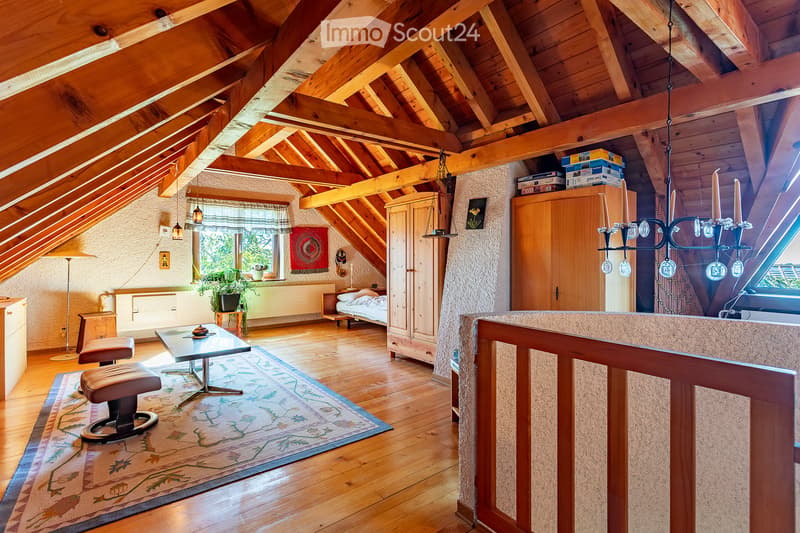 Cozy attic room with wooden beams, exposed wooden ceiling, and wooden floor. Features a table with two stools, a bed, a cabinet, a chandelier, and a plant near the window.