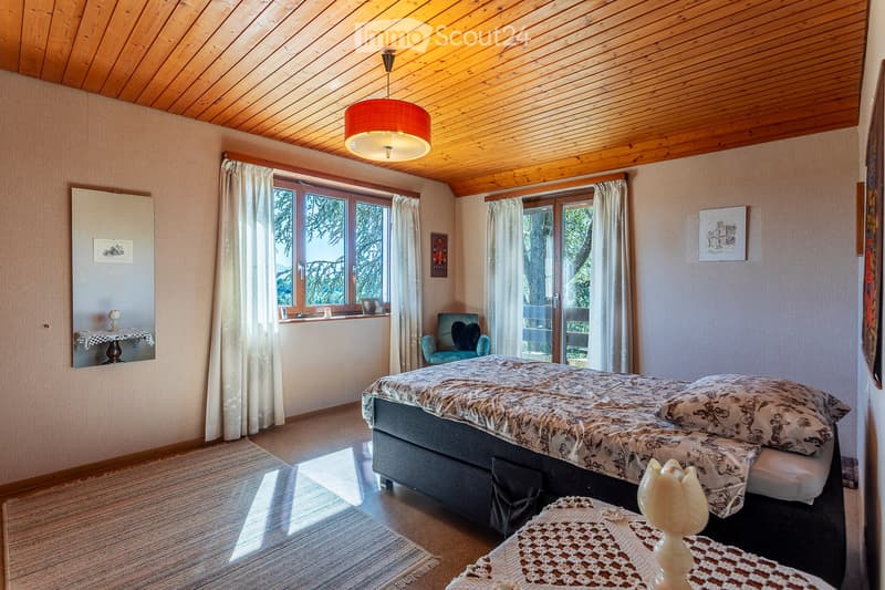 Cozy bedroom with wooden ceiling, large windows with curtains, a double bed with patterned bedding, a nightstand, a small table with a mirror, and a decorative chair near the window.