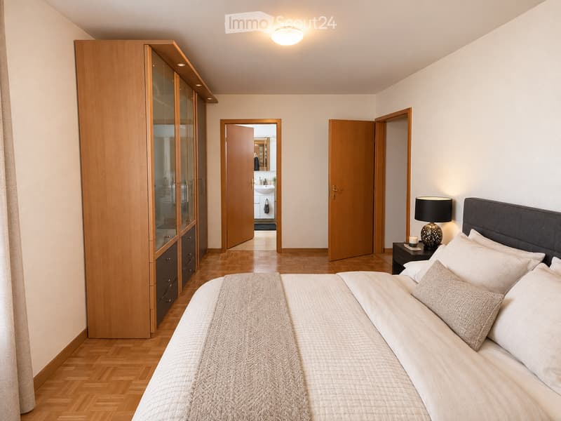 Modern bedroom with a large bed, nightstand, lamp, and a wooden wardrobe with mirrored doors. The room features light-colored walls, wooden flooring, and an open door leading to a bathroom.