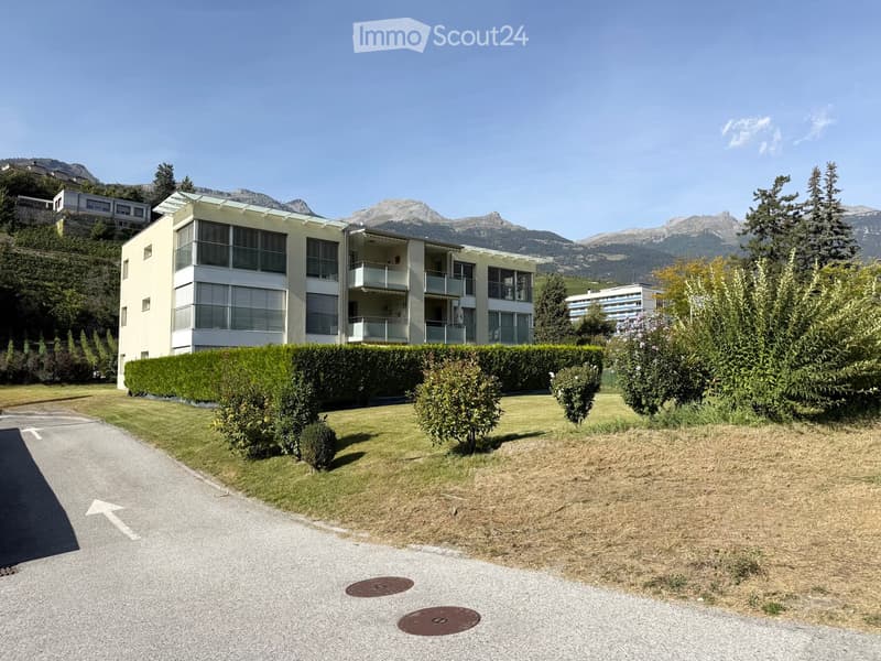 Modern multi-story building with balconies, surrounded by well-maintained hedges and trees, with a scenic mountain backdrop.