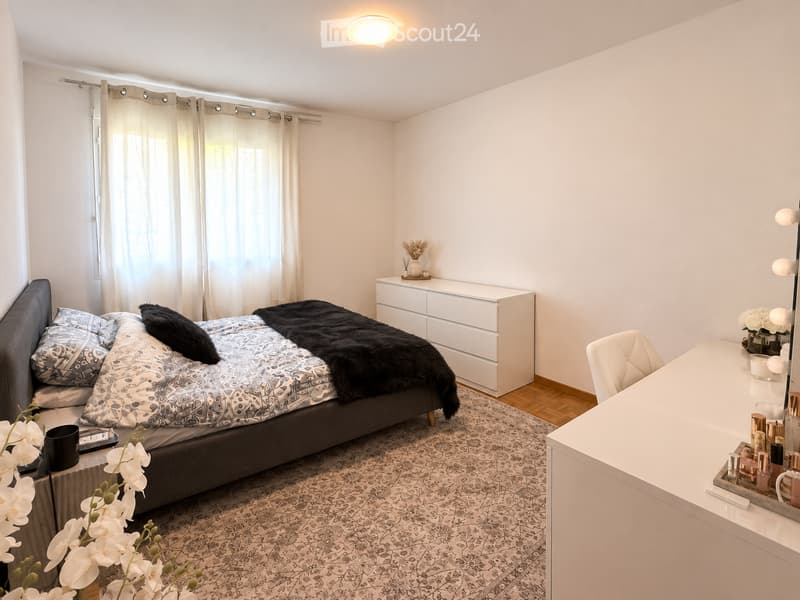 Modern bedroom with a large bed, patterned bedding, a nightstand with a vase, a vanity with a mirror, and a chair. The room features a patterned rug, white walls, and large windows with sheer curtains.