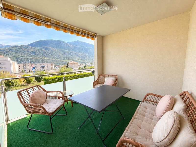 Balcony with green artificial turf, black rectangular table, beige wicker sofa, and two wicker chairs with pink cushions, overlooking a mountain view.